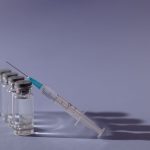 Seringa e frascos de vacina dispostos sobre uma superfície cinza.